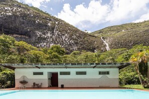 A pool area with a building that has a tiled facade and a door, surrounded by lush greenery and a mountainous backdrop. - S tio com Piscina e Lareira a 10 min de Itaipava (Petrópolis)