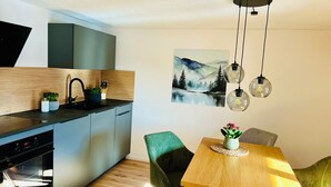 Uma cozinha moderna com forno embutido, uma área de jantar com mesa de madeira e cadeiras verdes, e um quadro de uma paisagem de montanhas na parede.