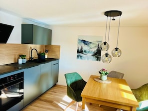 A modern kitchen with a built-in oven, a dining area with a wooden table and green chairs, and a painting of a mountain landscape on the wall. - Holiday Apartment 'Waldgeflüster' with Beautiful View, Private Balcony & Shared Garden (Ibach)