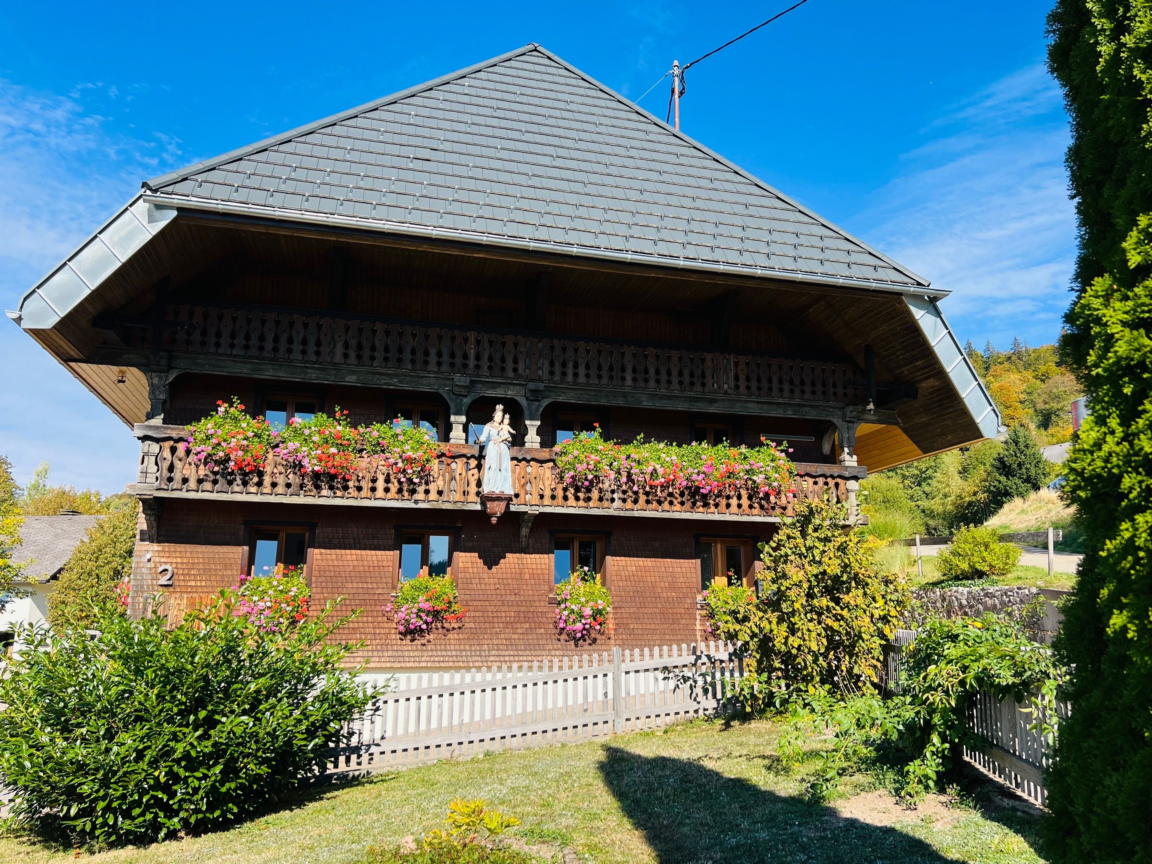 Ein traditionelles Chalet mit einem steilen Walmdach, Holzbalkonen mit Blumenkästen und einem weißen Zaun.