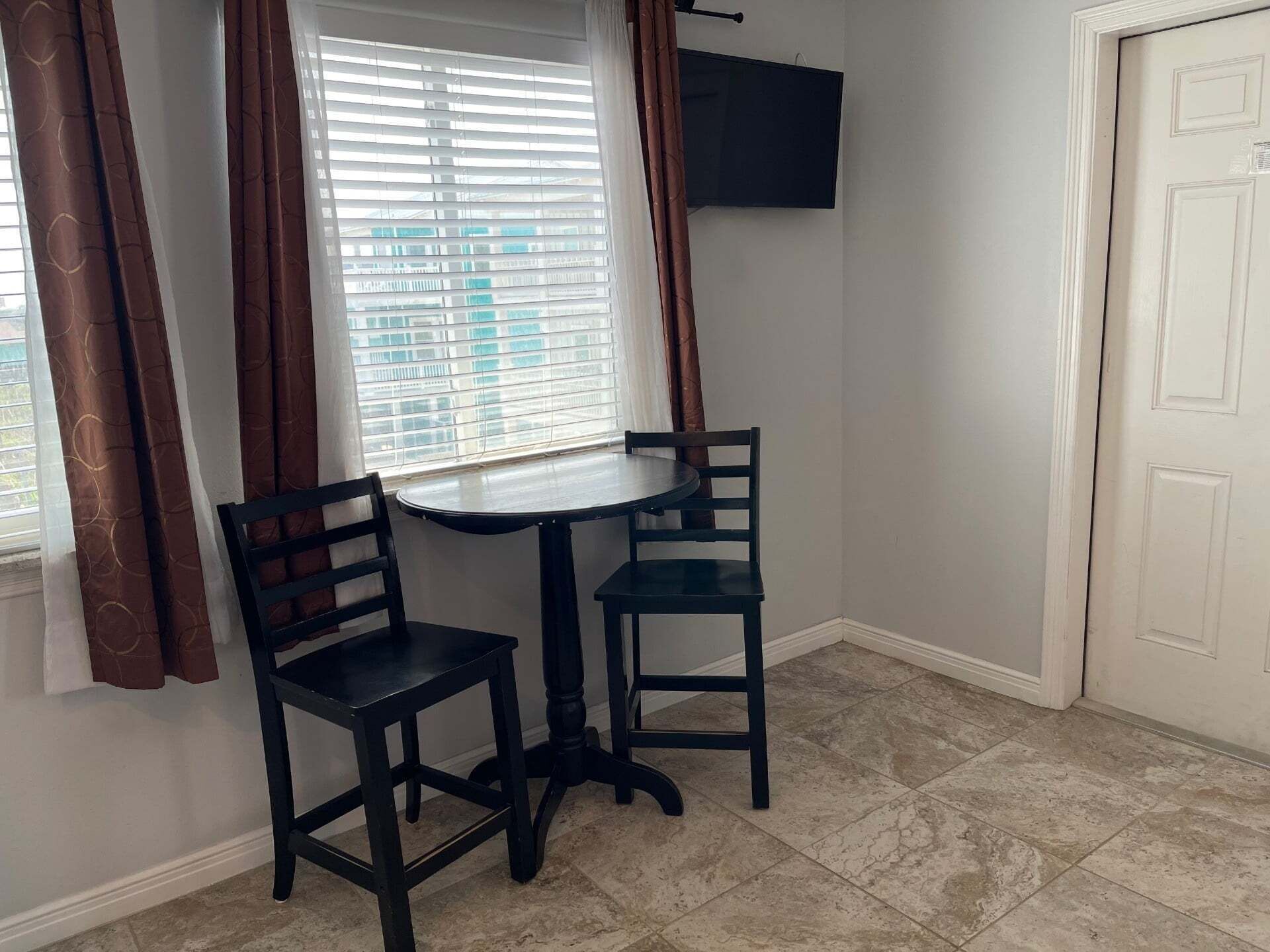 A corner room with a small table and two chairs, a window with blinds, a door, and a TV mounted on the wall.