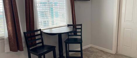 A corner room with a small table and two chairs, a window with blinds, a door, and a TV mounted on the wall.