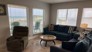 A living room with a sectional sofa, a single armchair, a coffee table, and large windows with blinds. - Beachgate Condo Suites And Hotel 237+239 2 Bedroom Condo (Port Aransas)