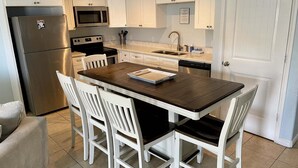 A kitchen with white cabinets, a stainless steel refrigerator, a microwave, a sink, and a dining table with four chairs. - Beachgate Condo Suites And Hotel 536+537b 2 Bedroom Condo (Port Aransas)
