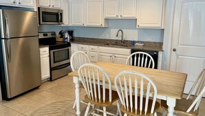 A kitchen with white cabinets, a stainless steel refrigerator, a microwave, an oven, a sink, and a dining table with four chairs. - Beachgate Condo Suites And Hotel 526+527b 2 Bedroom Condo (Port Aransas)