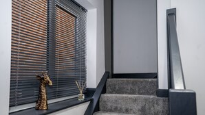 A staircase with grey carpeted steps and a black handrail, leading to a room with white walls and a window with black blinds. - Rose House (Boston)