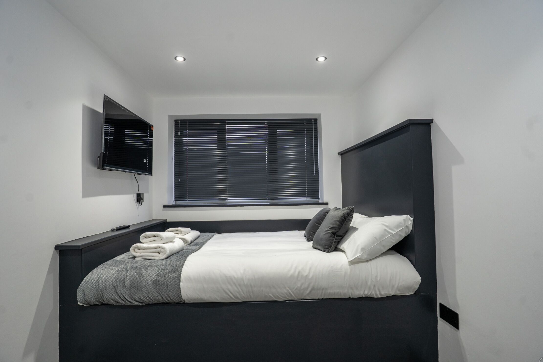 Un dormitorio moderno con una cama, un televisor montado en la pared y una ventana con persianas.