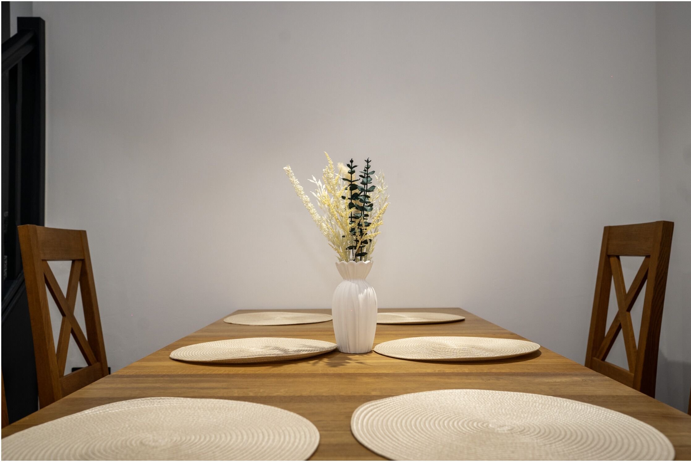 Una mesa de comedor con un jarrón blanco que contiene flores secas, rodeada de individuales tejidos.