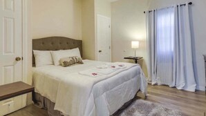 Un dormitorio ordenado con una cama grande, una mesita de noche, una lámpara y una ventana con cortinas.
