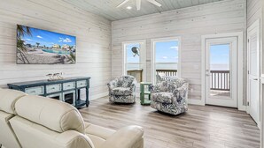 Ruime kamer met plafondventilator, groot raam met uitzicht op het strand, een bank en twee stoelen.
