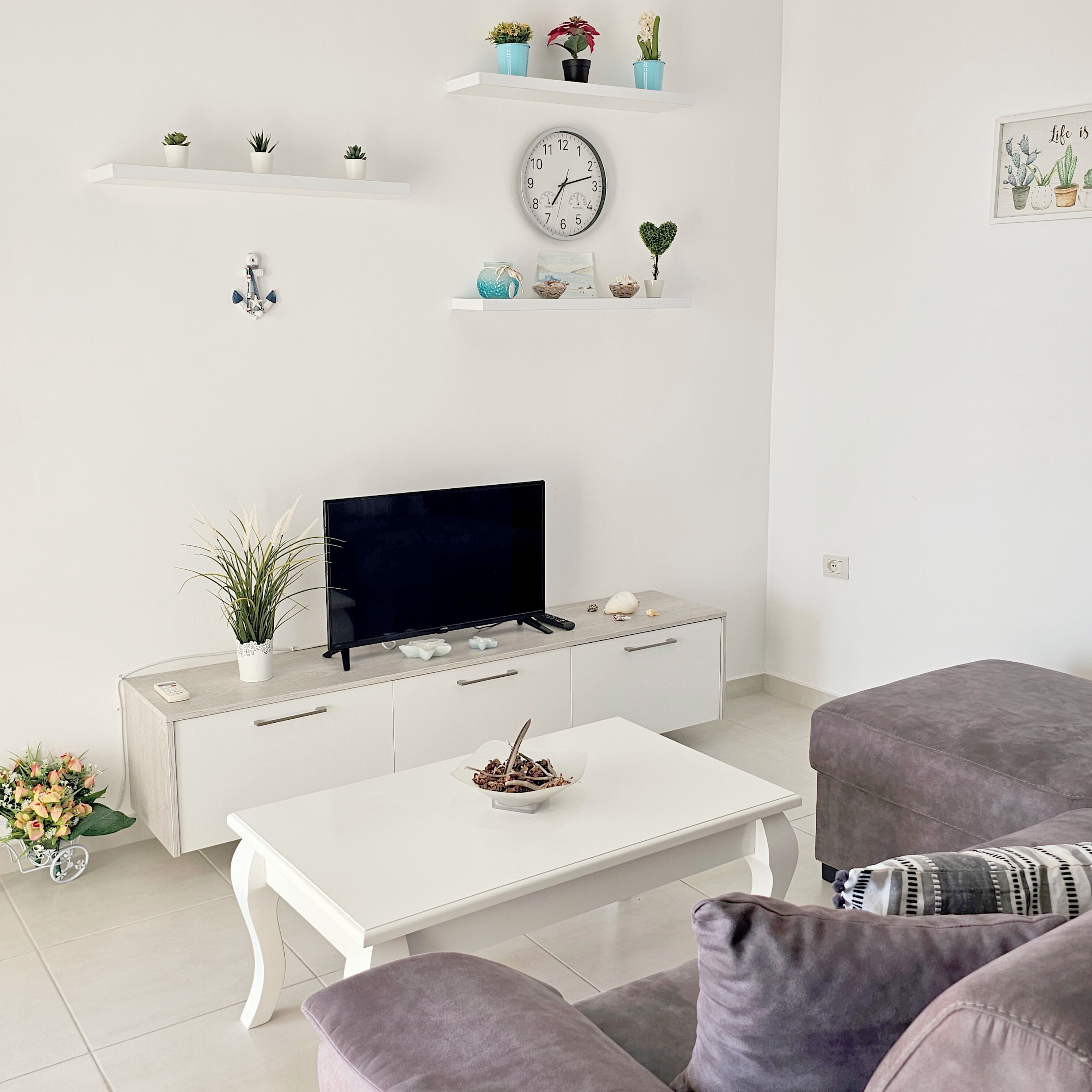 Una sala de estar moderna con un televisor de pantalla plana sobre un mueble blanco, una mesa de centro, dos sofás grises y objetos decorativos en estanterías.