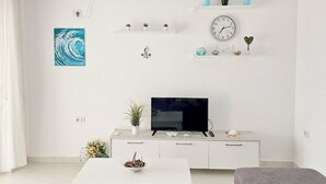 Ein modernes Wohnzimmer mit Flachbildfernseher, weißem Sofa und Couchtisch. An den Regalen stehen Pflanzen und an der Wand hängt eine Uhr.