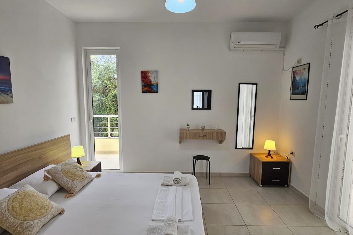 A hotel room with a bed, bedside lamps, a small table, a mirror, and a view of the outdoors.