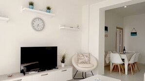 Ein modernes Wohnzimmer mit Flachbildfernseher, weißen Sofas, einem Couchtisch und einem Essbereich mit Tisch und Stühlen.