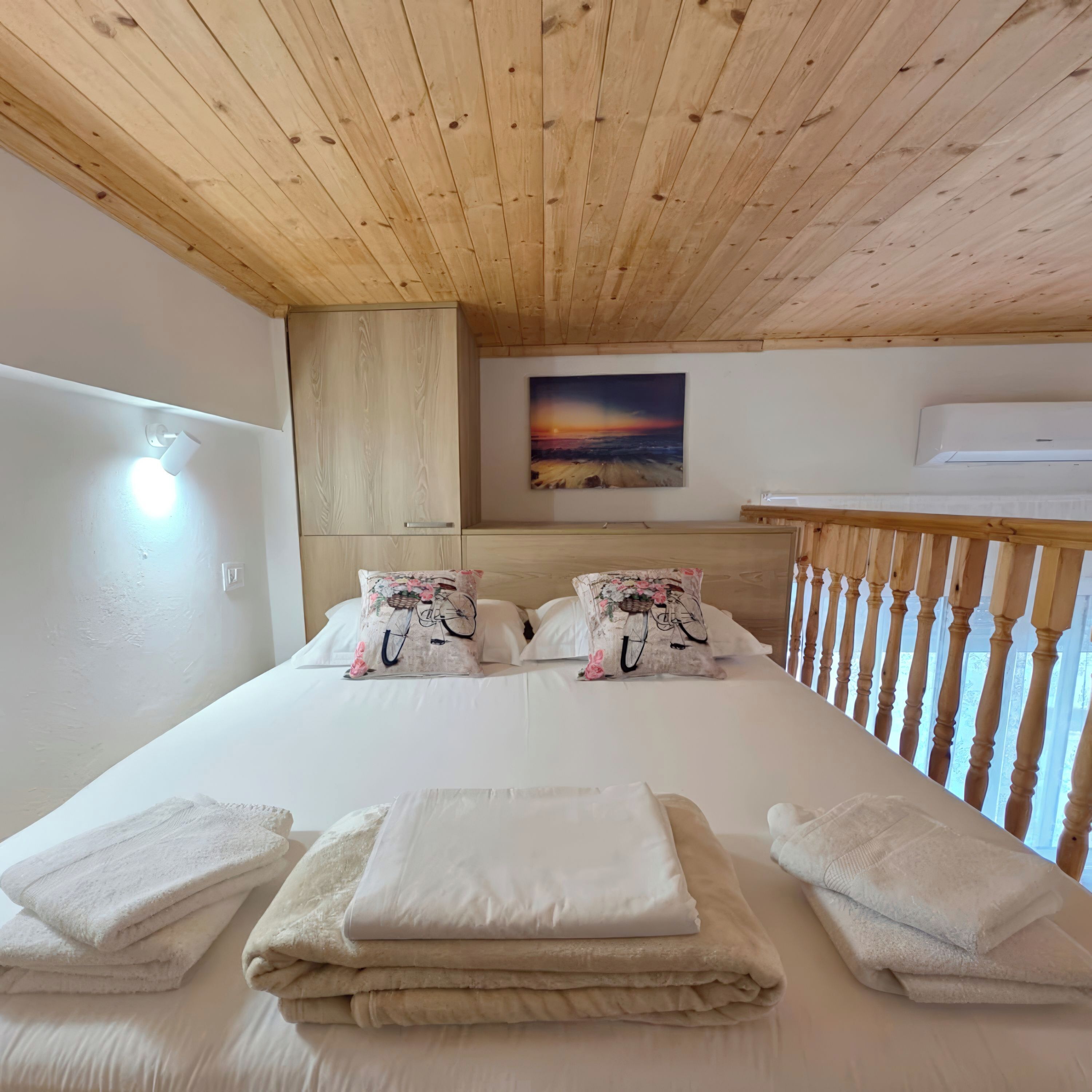 A bedroom with a wooden ceiling, a bed with white linens, two pillows with floral designs, a painting on the wall, and an air conditioning unit.