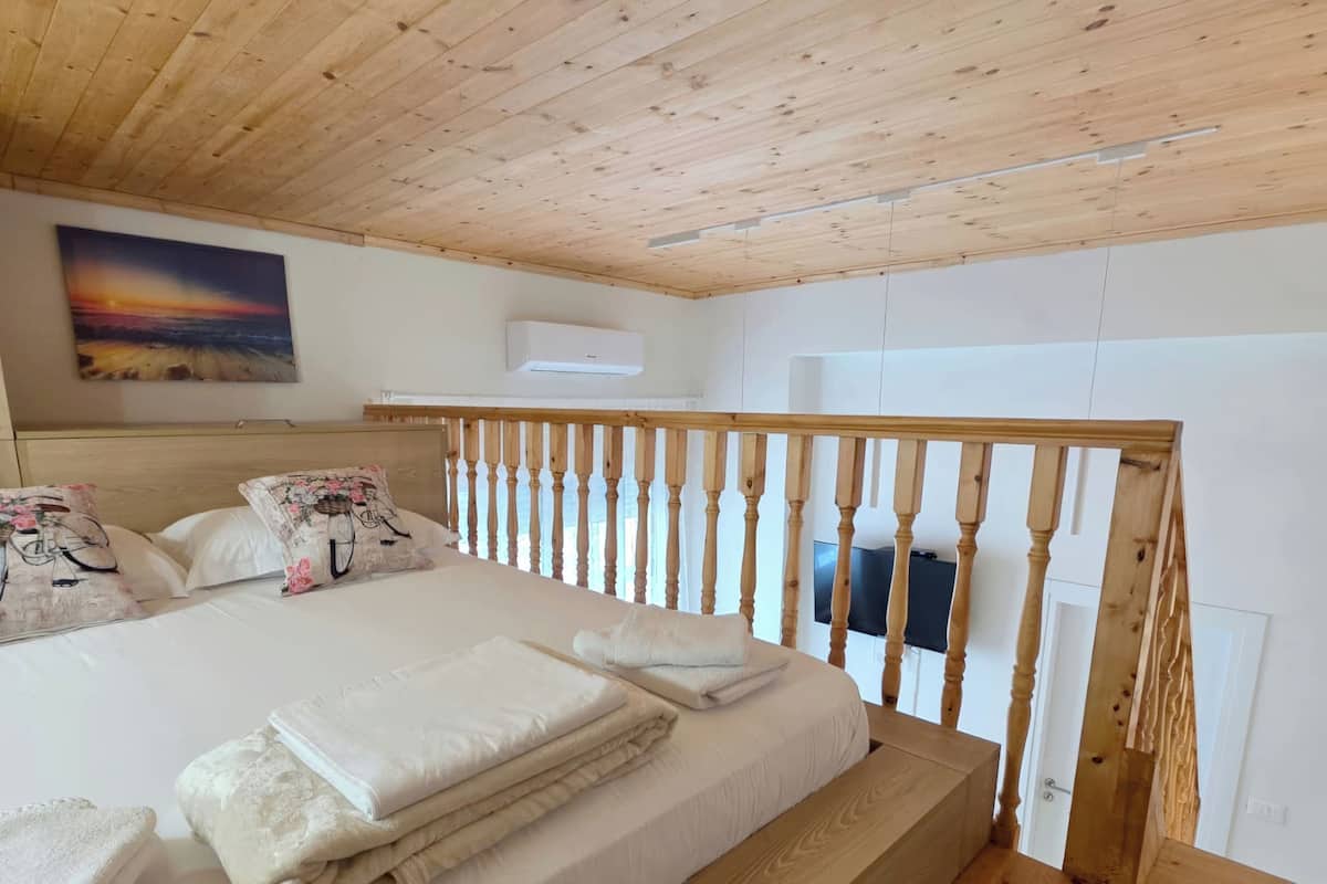 A bedroom with a wooden ceiling, a bed with white bedding, a painting on the wall, and a TV mounted on the wall.