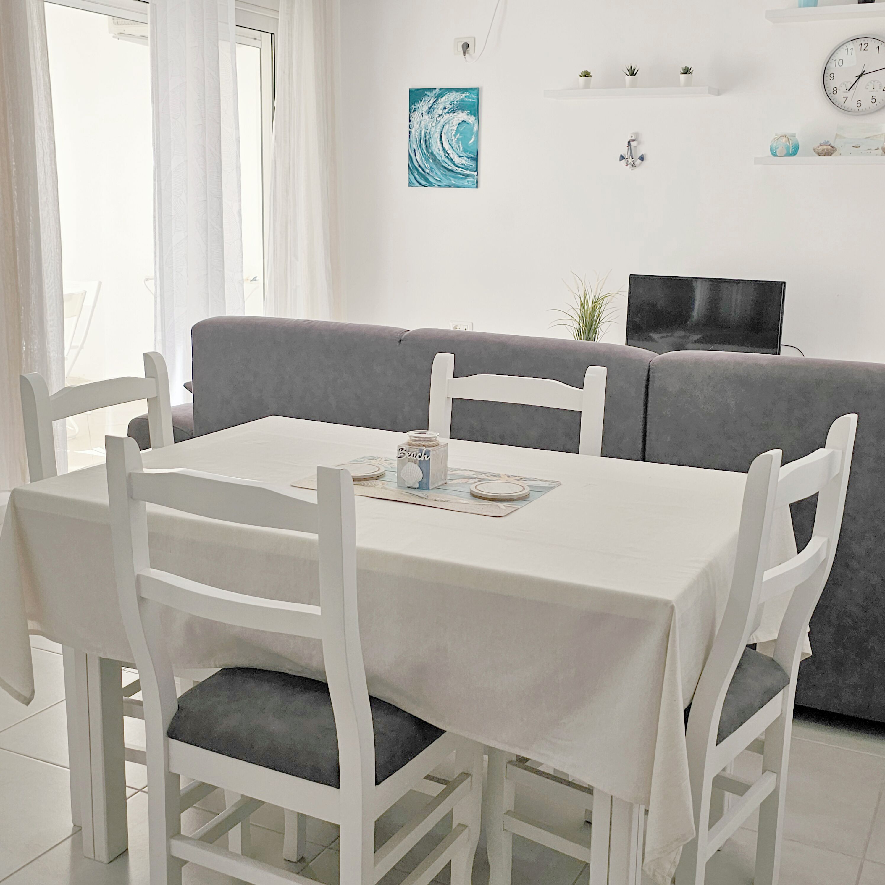 Een eetgedeelte met een witte tafel en stoelen, een grijze bank en een wandklok.