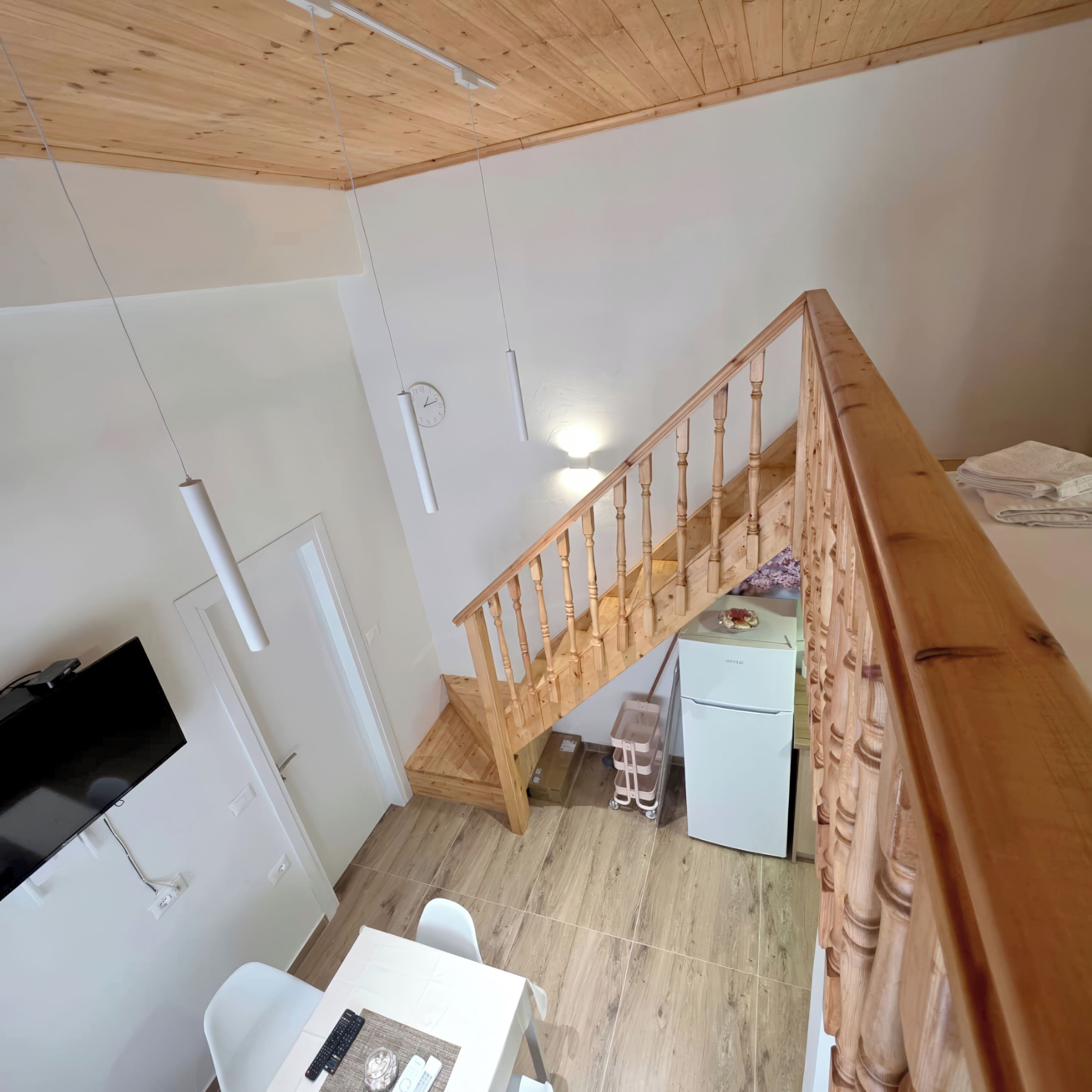 A loft-style room with a wooden staircase, a white refrigerator, a small kitchen area, and a white desk with a chair.