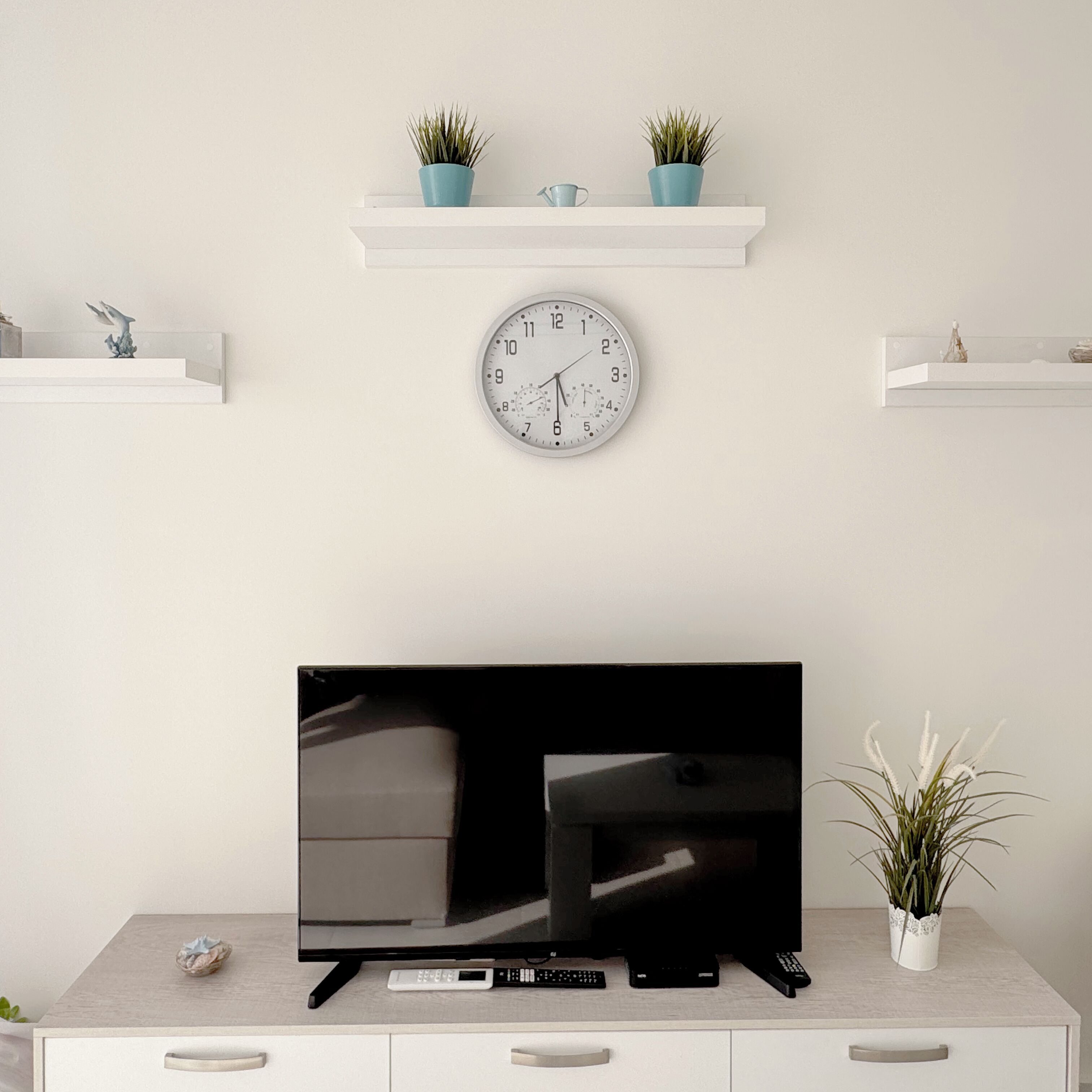 A modern living room with a flat-screen TV on a wooden cabinet, a wall clock, and two shelves with potted plants.