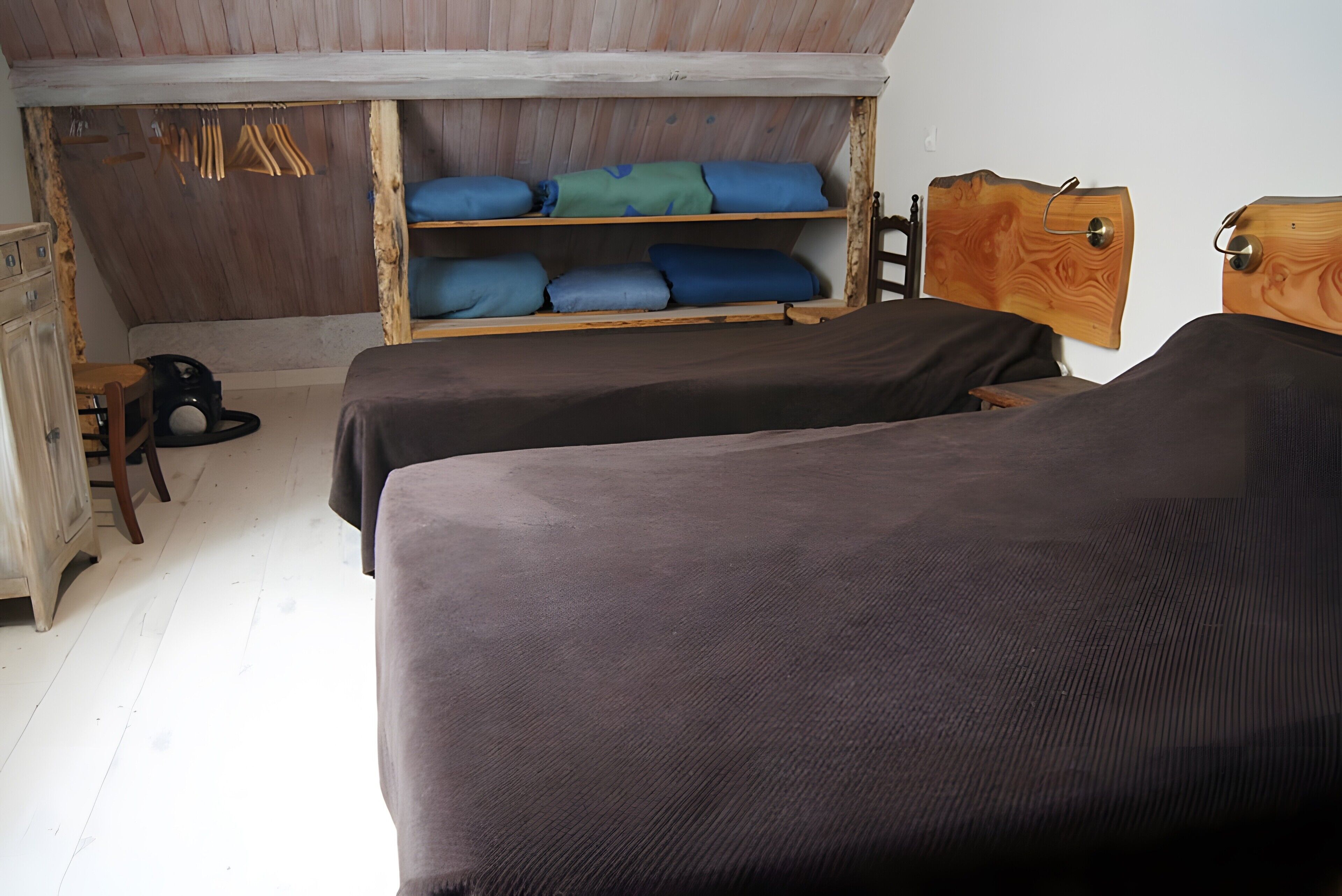 A room with two beds, wooden headboards, a wardrobe, and a shelf with folded clothes.