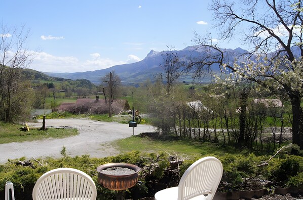A patio with two white chairs, a small fire pit, and a view of a rural landscape with mountains and a house in the distance.