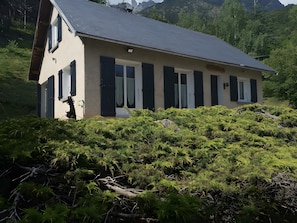 A single-story house with a sloped roof, surrounded by dense green vegetation and mountains in the background.
