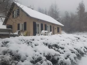 En snötäckt byggnad med ett sluttande tak, en uteplats med utemöbler och snötyngda buskar.