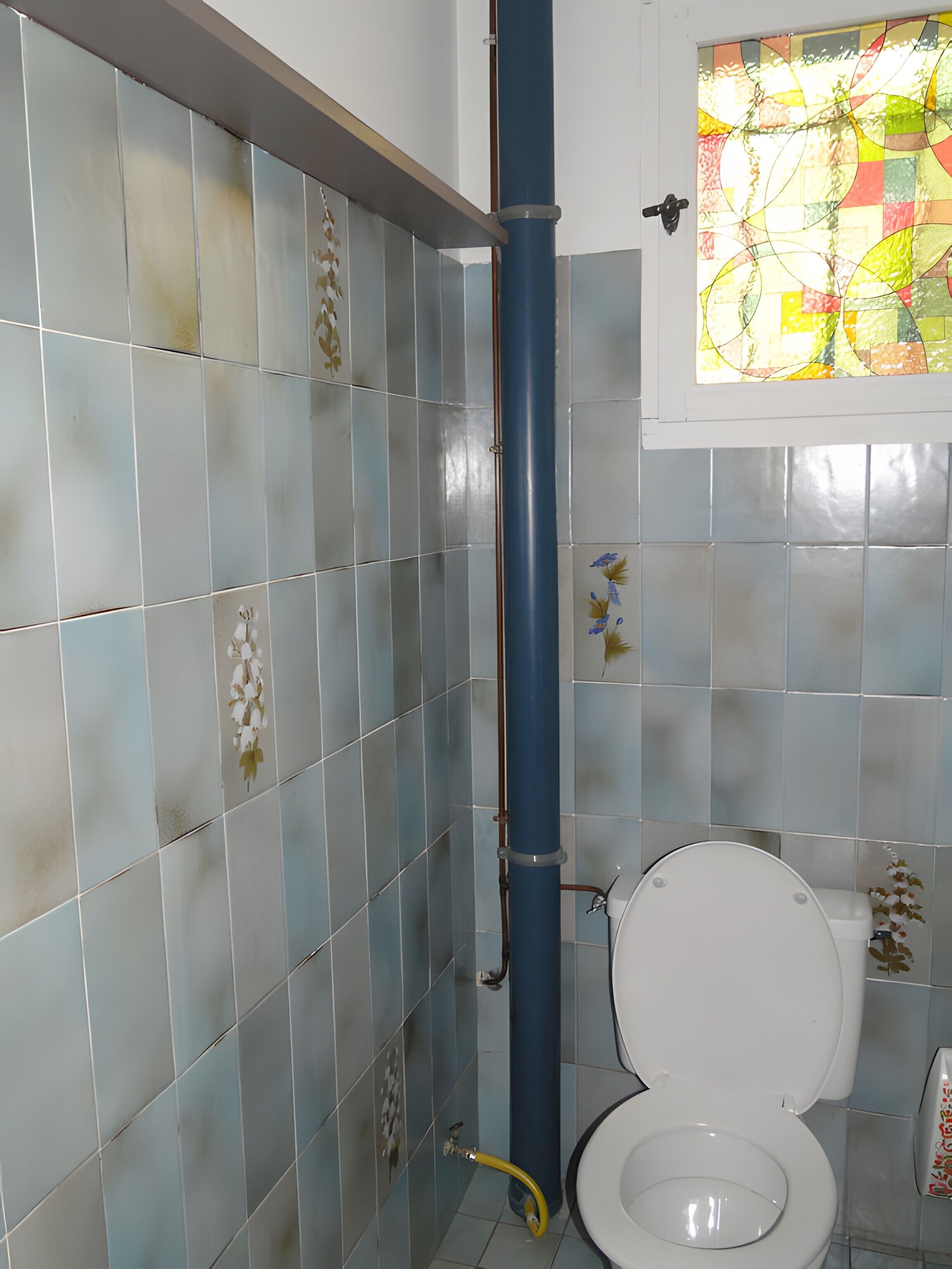 Un bagno con pareti piastrellate, un water, un tubo blu e una finestra con vetrate colorate.