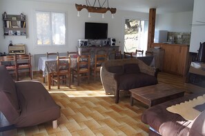 A living room with a sofa, chairs, a dining table, a television, and a kitchen area.