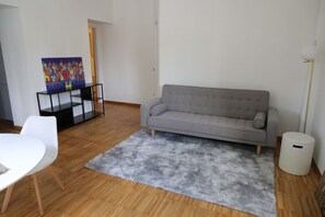 Een woonkamer met een grijze bank, witte stoelen, een grijs vloerkleed, een kleurrijk abstract schilderij en een hoge staande lamp.