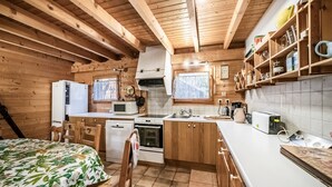 A wooden cabin interior with a kitchen, dining area, and a bed with a floral bedspread. - Chalet “8p Simple - Les Mésanges” with Mountain View, Private Terrace, and Wi-Fi (Verchaix)