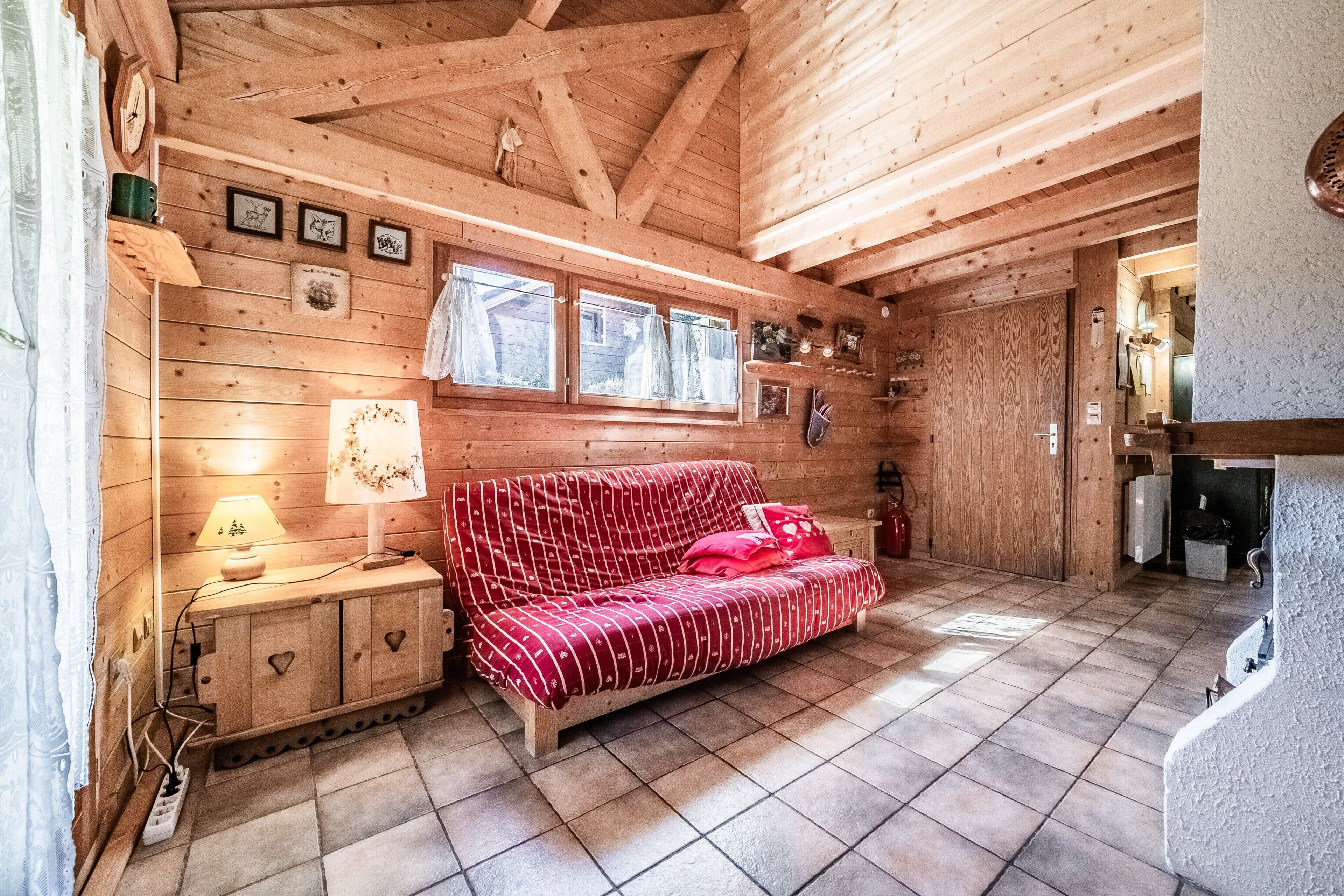 Interno di una baita in legno con un divano rosso a motivi decorativi, pareti in legno, pavimento piastrellato e una zona cucina visibile attraverso una porta aperta.