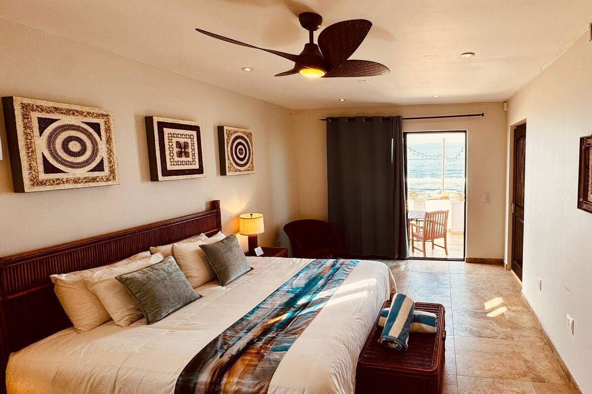 Een hotelkamer met een groot bed, een plafondventilator en uitzicht op de oceaan door een schuifdeur.