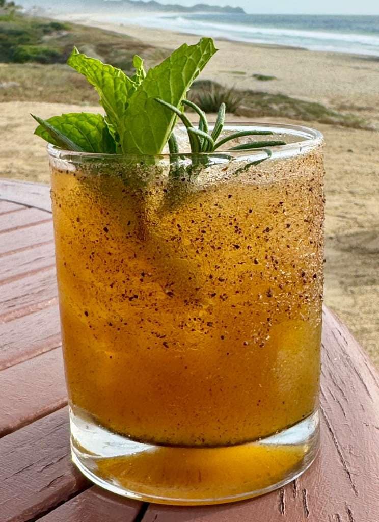 Un vaso de té helado con hojas de menta sobre una mesa de madera junto a la playa.