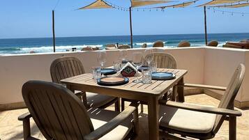 Zona pranzo all'aperto con tavolo imbandito per due persone, sedie e vista sull'oceano.