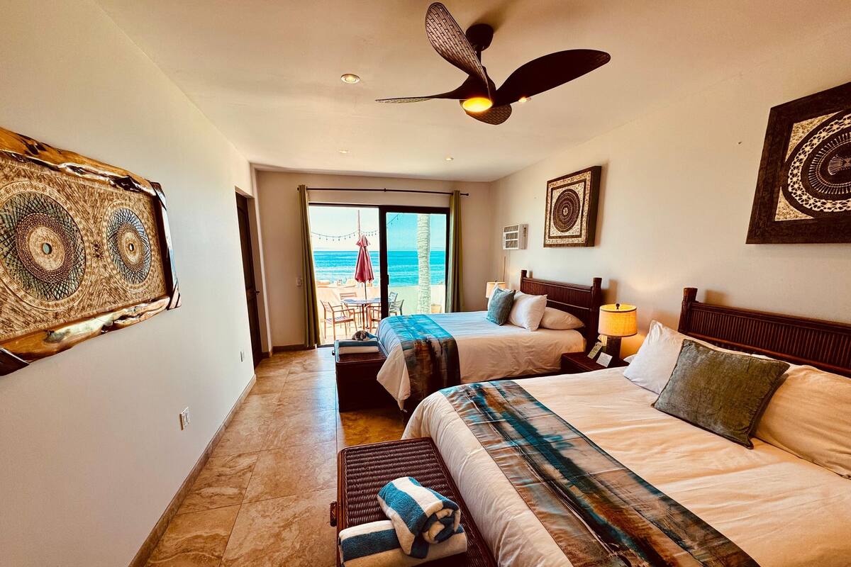 Een hotelkamer met twee bedden, een plafondventilator, uitzicht op het strand en een balkon met buitenmeubels.