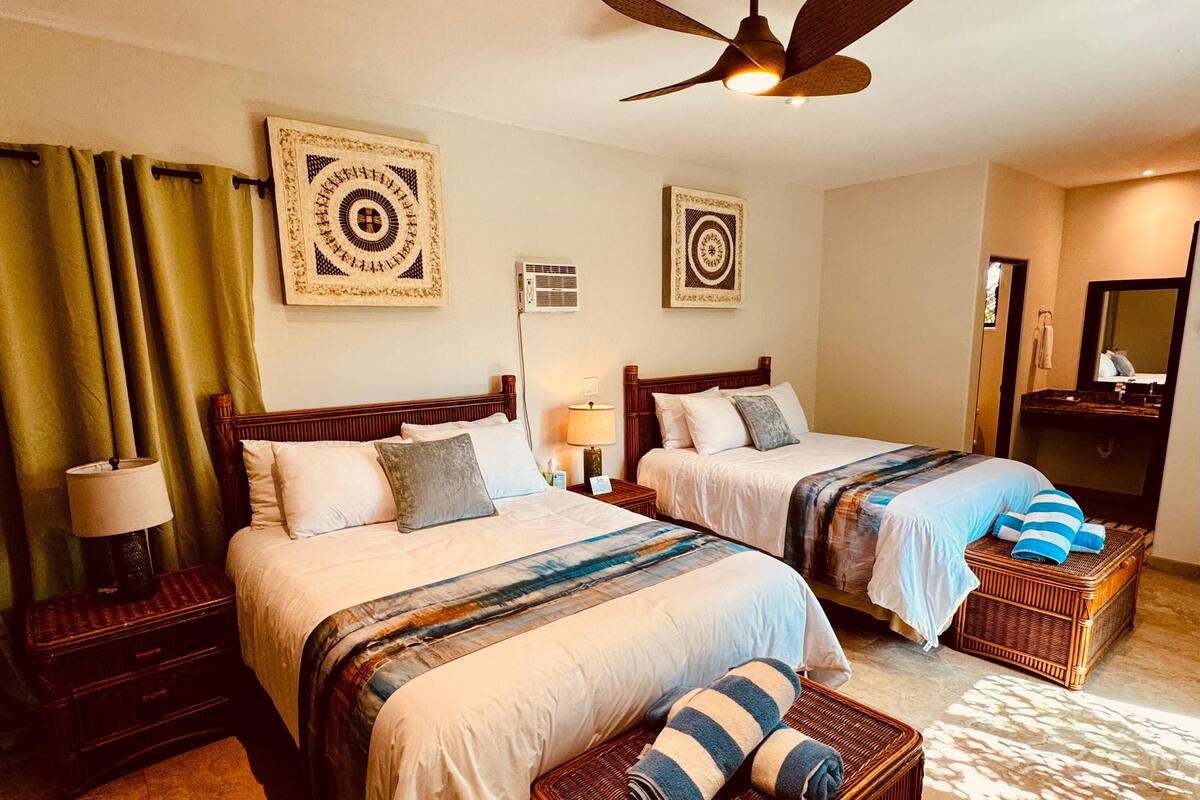 Een hotelkamer met twee bedden, een plafondventilator en een badkamer met spiegel en wastafel.