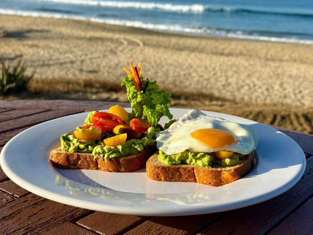 Une assiette de tartines à l’avocat, surmontées d’un œuf au plat, posée sur une table en bois, près de la plage.