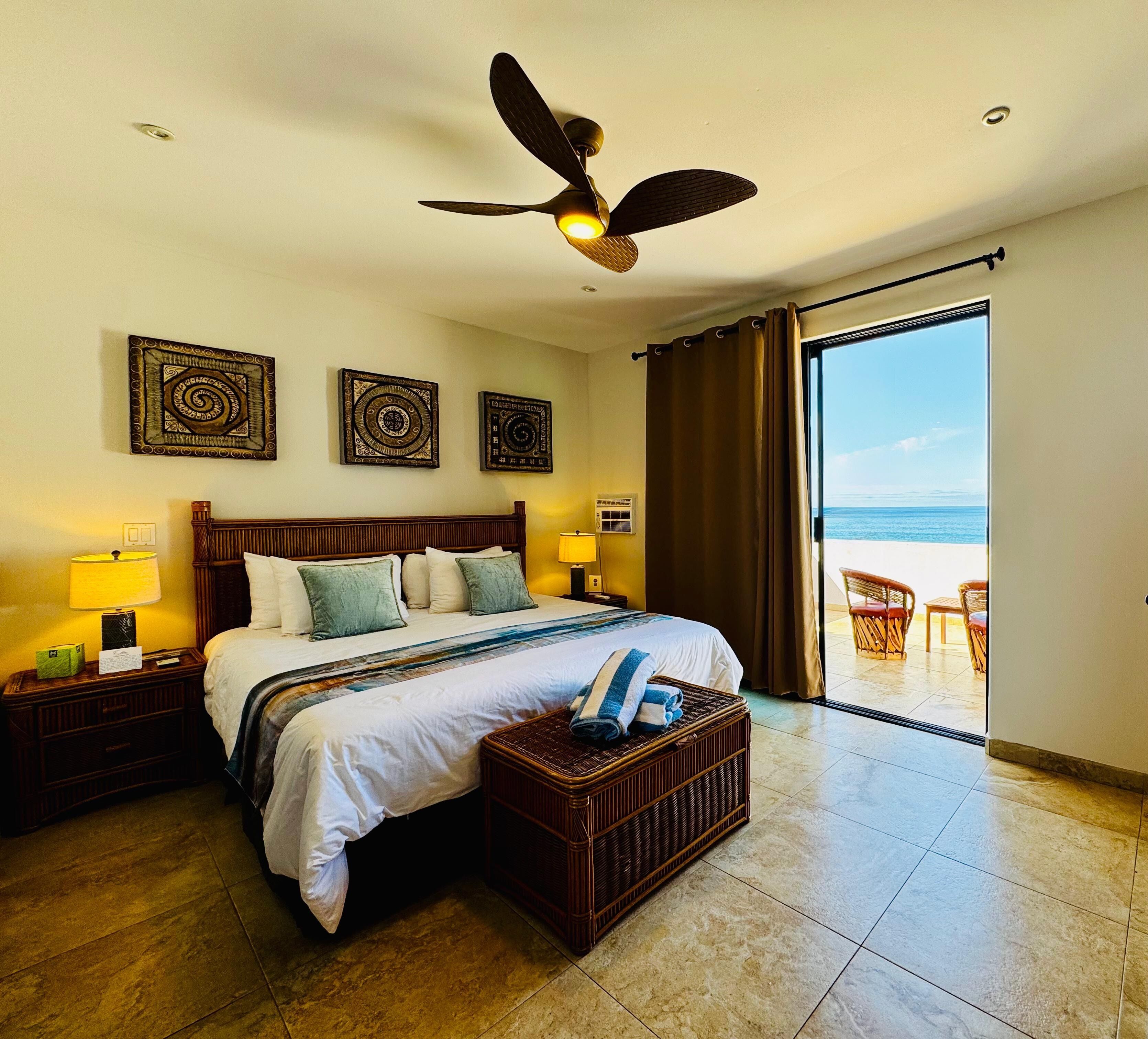 A hotel room with a bed, a ceiling fan, a view of the beach, and two wall art pieces.