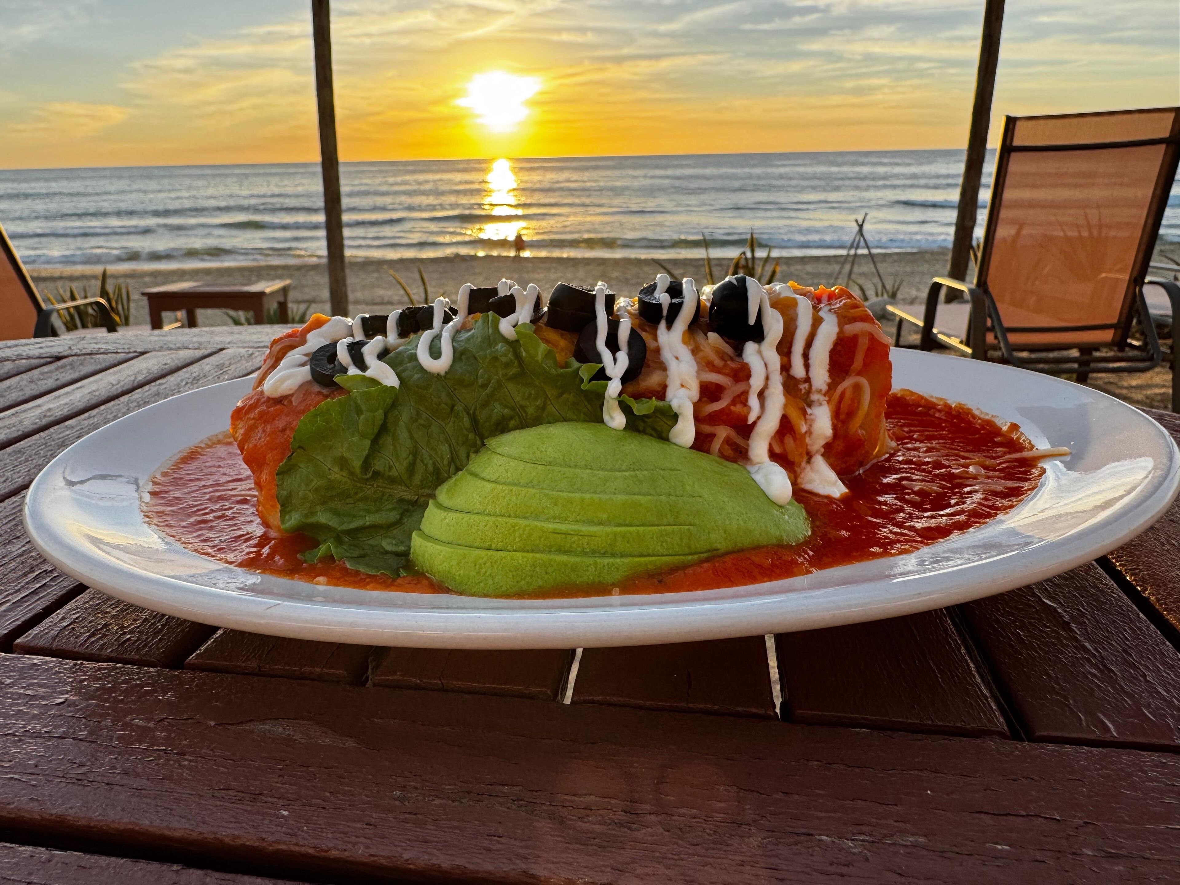 Une assiette d’enchiladas garnies, posée sur une table en bois, avec vue sur le coucher de soleil sur l’océan.