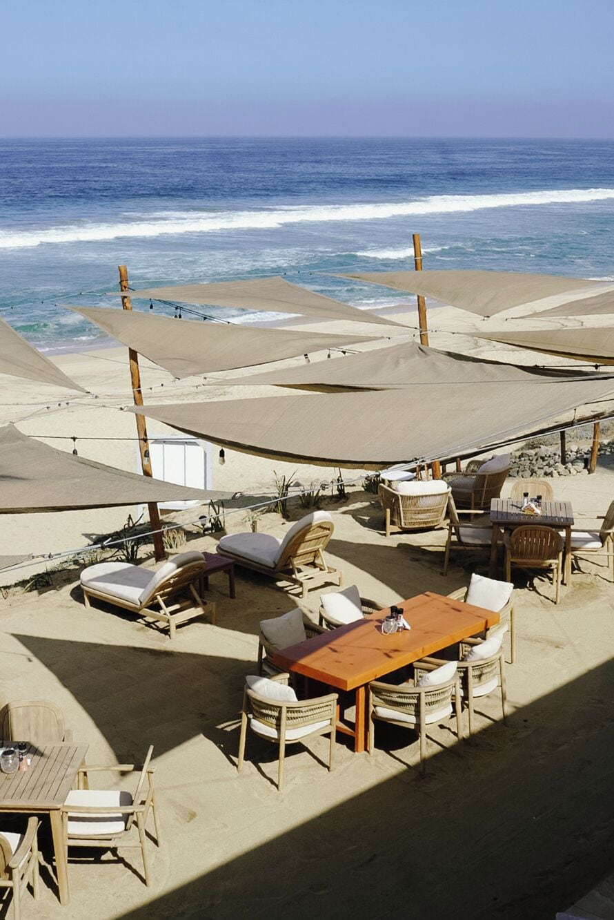 A beachside outdoor seating area with tables and chairs, shaded by large canopies, overlooking the ocean.