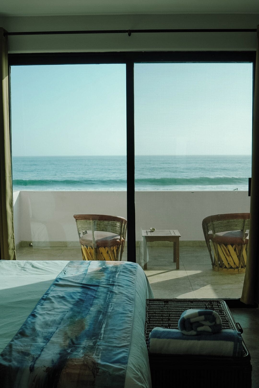 Habitación de hotel con cama, una mesita pequeña, dos sillas de mimbre y vista al océano.