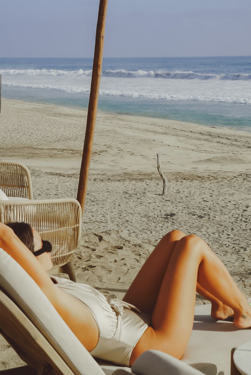 Escena de playa con una persona recostada en una silla de playa, un poste de madera y el océano al fondo.