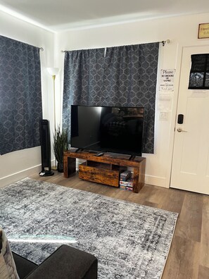 Una sala de estar con un televisor de pantalla plana sobre un soporte de madera, cortinas estampadas, una planta en maceta y un cartel de 'No Fumar' en la puerta.