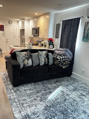 Una sala moderna con un sofá negro, alfombra estampada, cocina al fondo y un reloj en la pared.