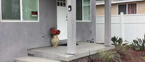 A modern two-story house with a covered front porch, a front door, and a small garden area with plants and a flower pot.