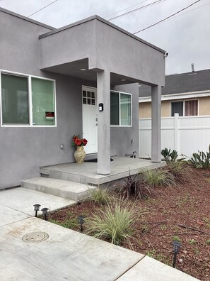 Une maison moderne à deux étages, dotée d’un porche avant couvert, d’une porte d’entrée et d’un petit jardin agrémenté de plantes et d’un pot de fleurs.