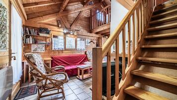 A wooden cabin interior with a staircase, a red sofa, a wooden chair, and a bookshelf.