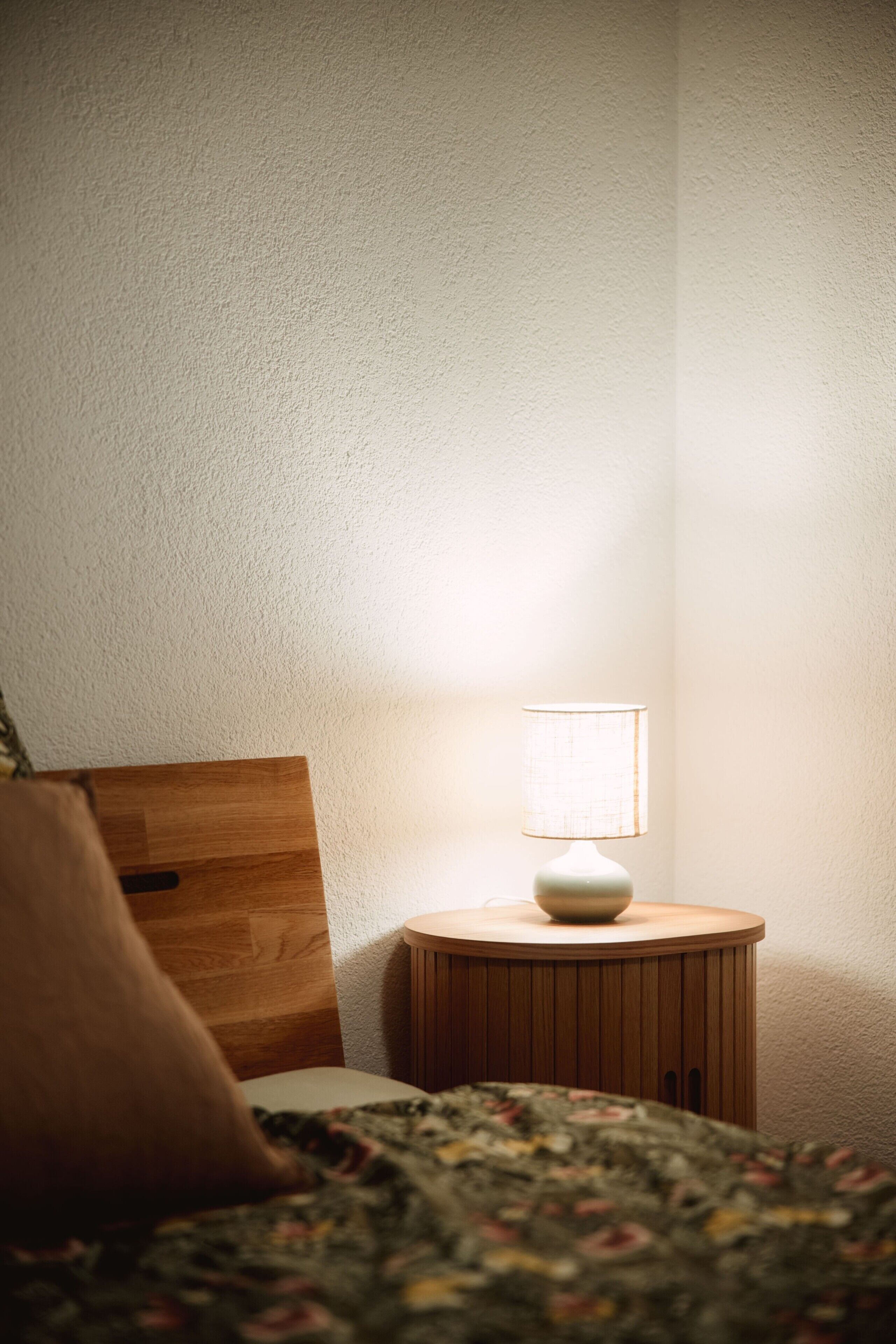 Une chambre à coucher avec une tête de lit en bois, une table de chevet ronde surmontée d’une lampe, et une couvre-lit à motifs floraux.