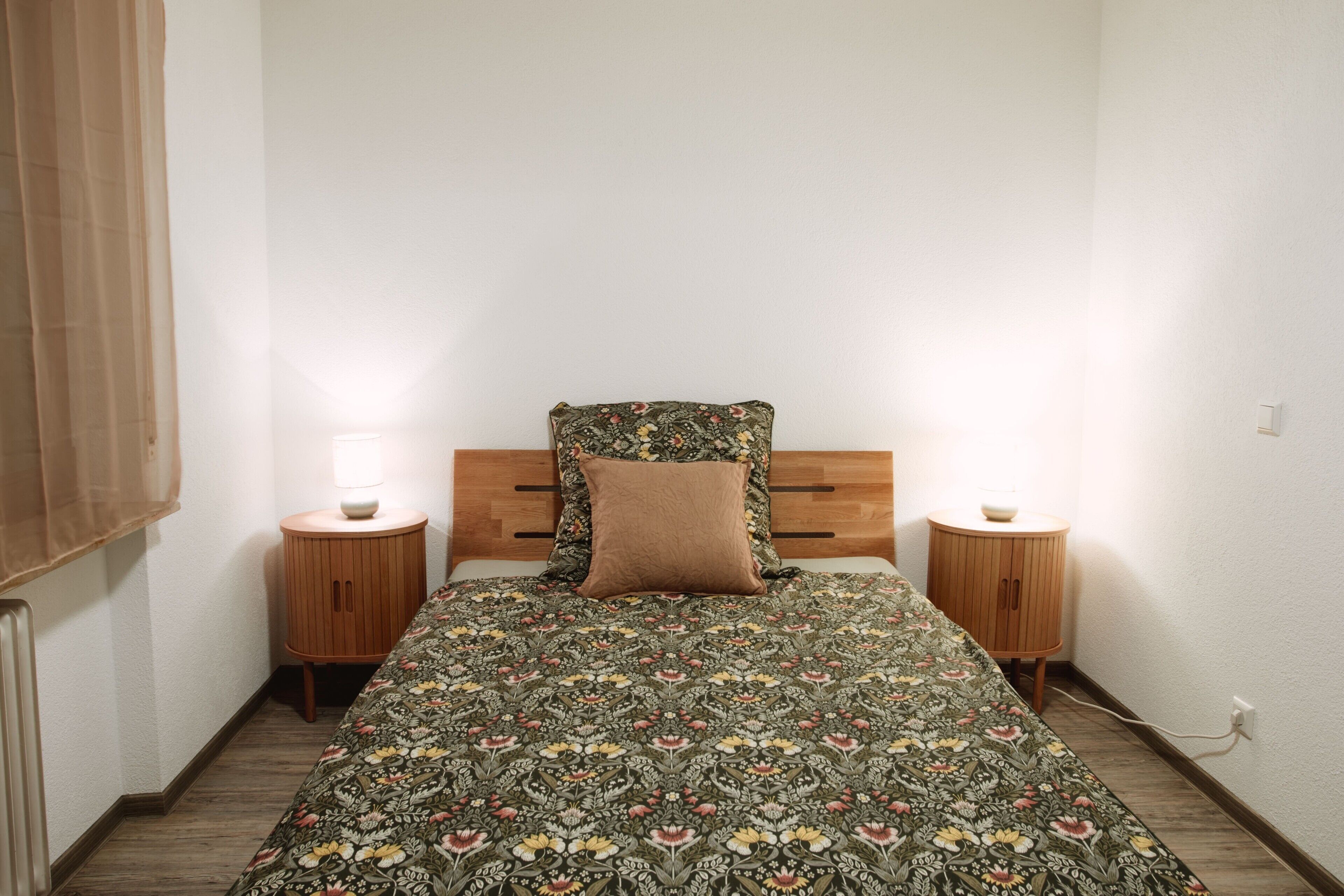 A bedroom with a wooden headboard, a floral bedspread, two bedside tables with lamps, and a wooden floor.
